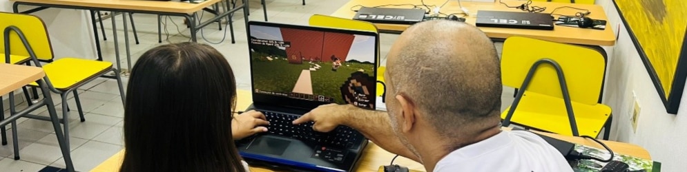 Educator points to a laptop screen while helping a student use Minecraft in a classroom.