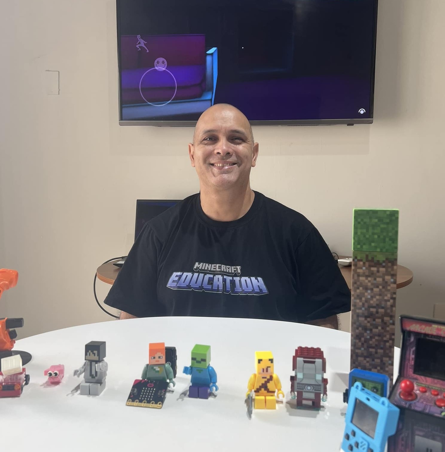 Educator in a Minecraft Education shirt sits behind a table with Minecraft figures, a small arcade machine, and block toys.