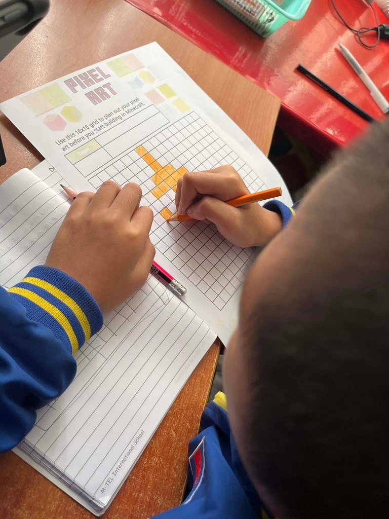 A student uses an orange pencil to draw pixel art on a 16x16 grid planning worksheet at their desk.