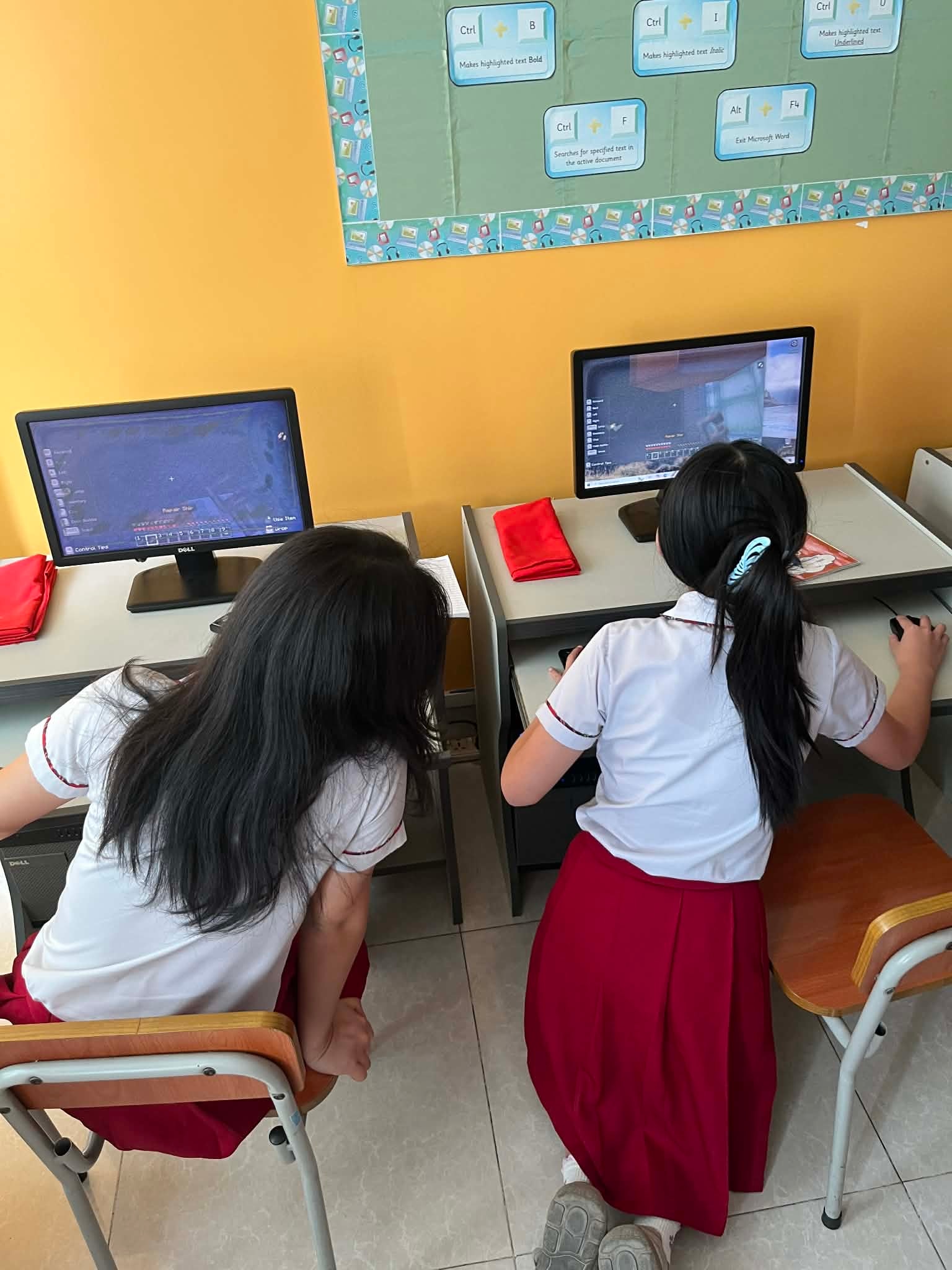 Two students in school uniforms play on desktop computers in a classroom.