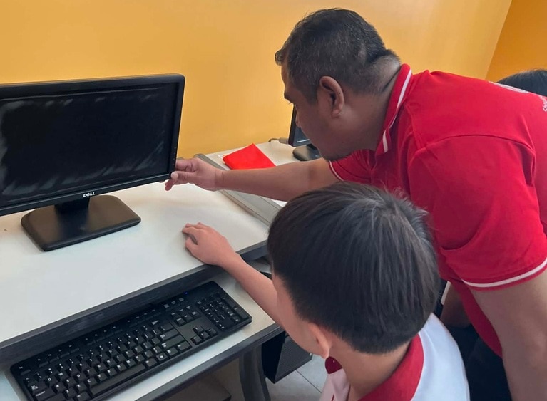 A teacher points at a desktop computer screen while helping a student in a classroom.