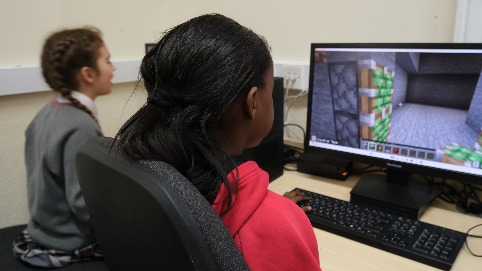A student plays Minecraft Education, exploring a stone structure, as a classmate sits beside her.