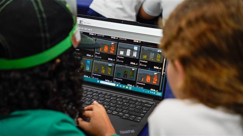 Two students look at a laptop screen showing a Minecraft server or world selection menu.