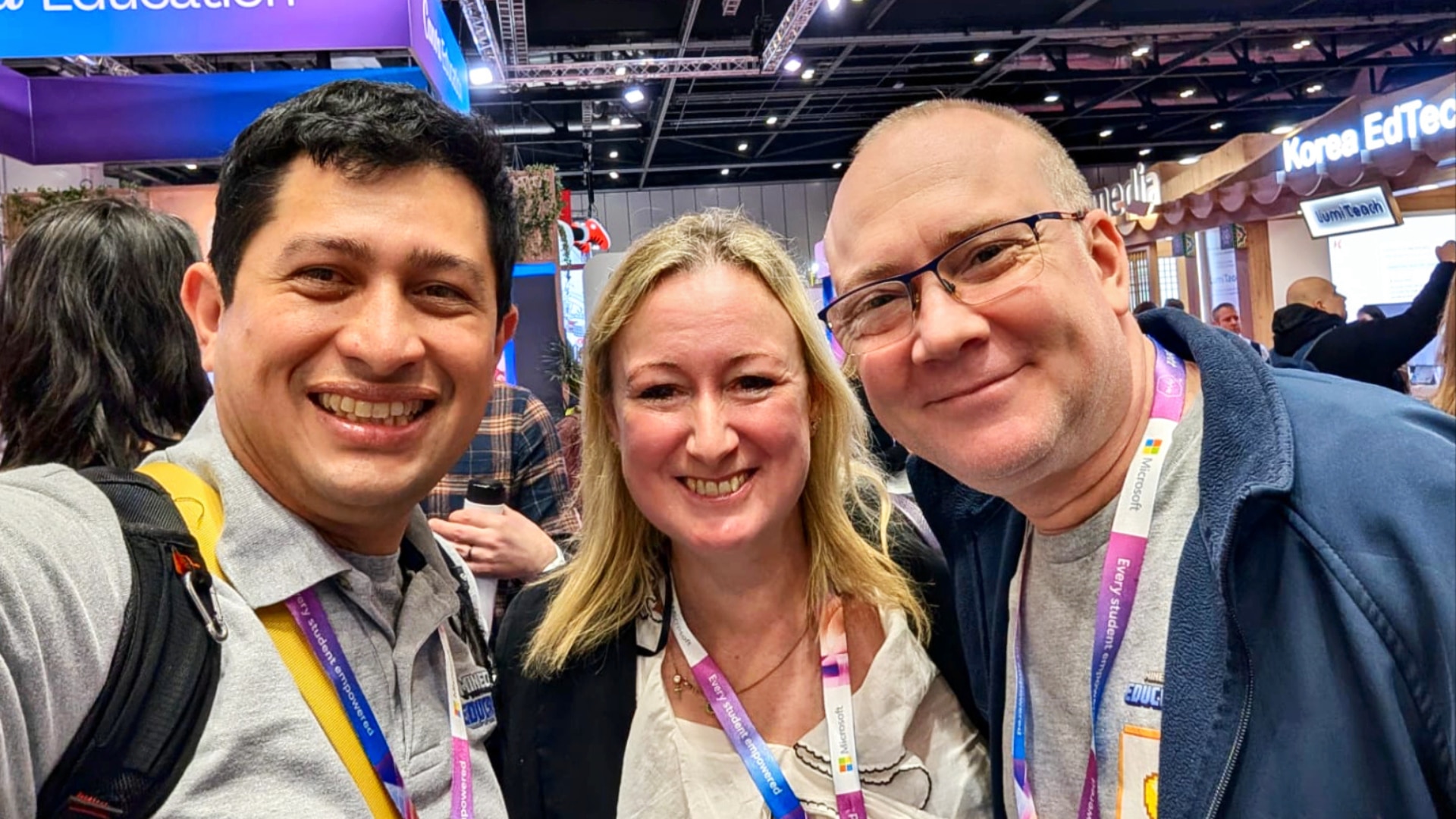 A selfie of three smiling attendees, two men and one woman wearing event lanyards, in a busy Microsoft Education exhibition hall.