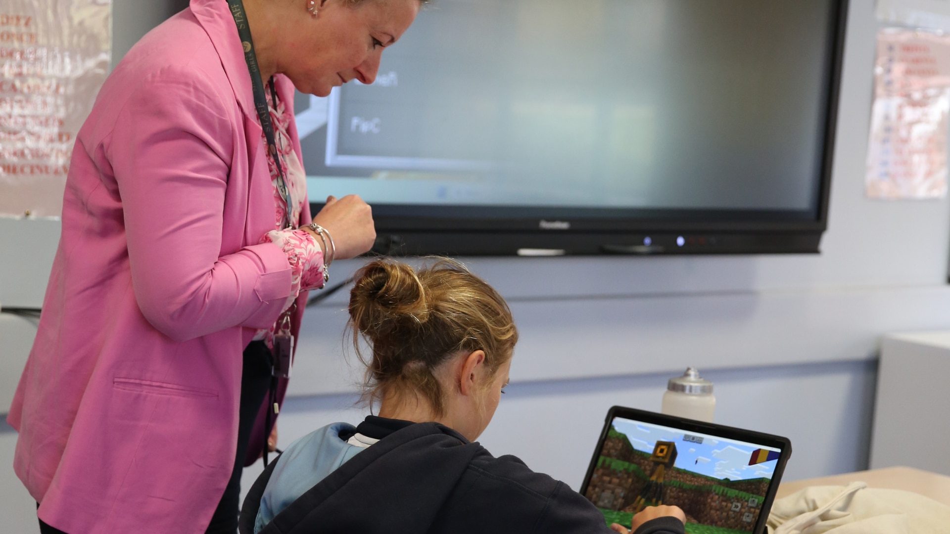 A teacher in a pink blazer leans over to help a young student playing Minecraft on a laptop in a classroom, with a large display screen on the wall behind them.
