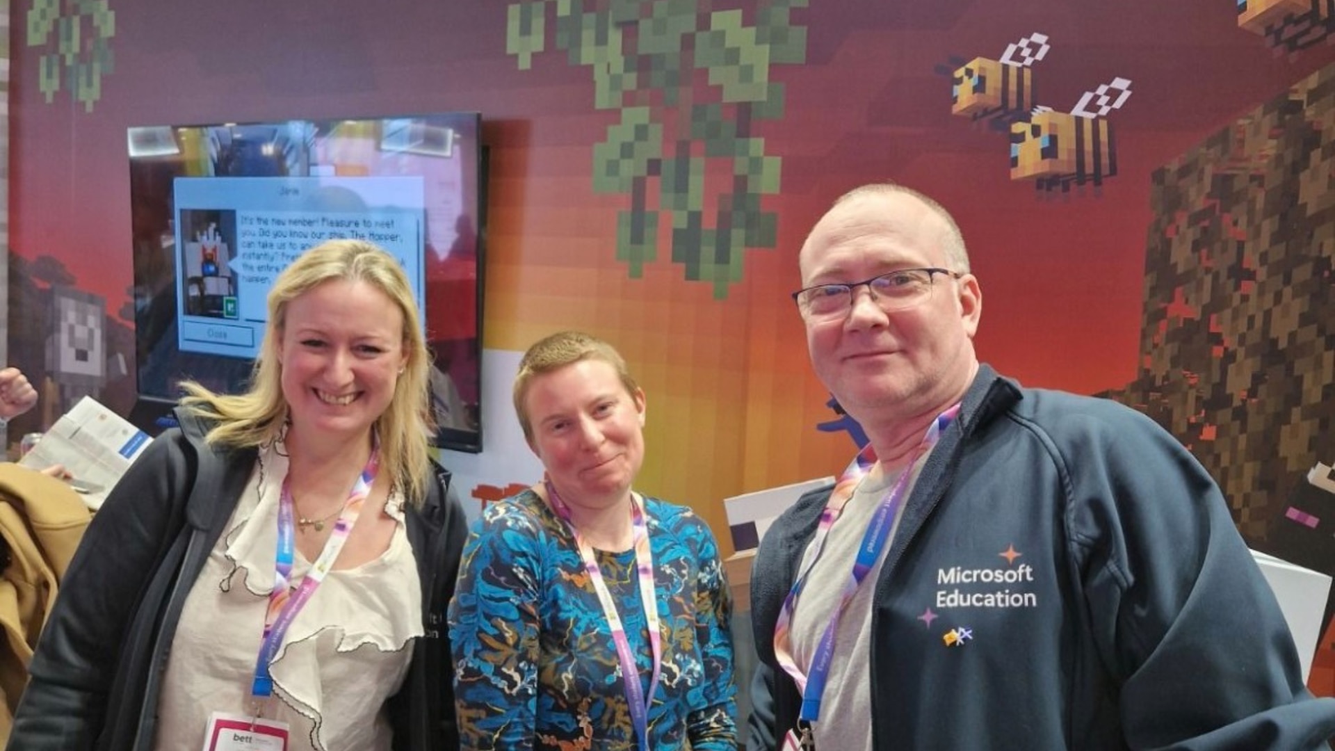 Three attendees, one wearing a Microsoft Education jacket, pose in front of a Minecraft display featuring pixelated bees and vines.