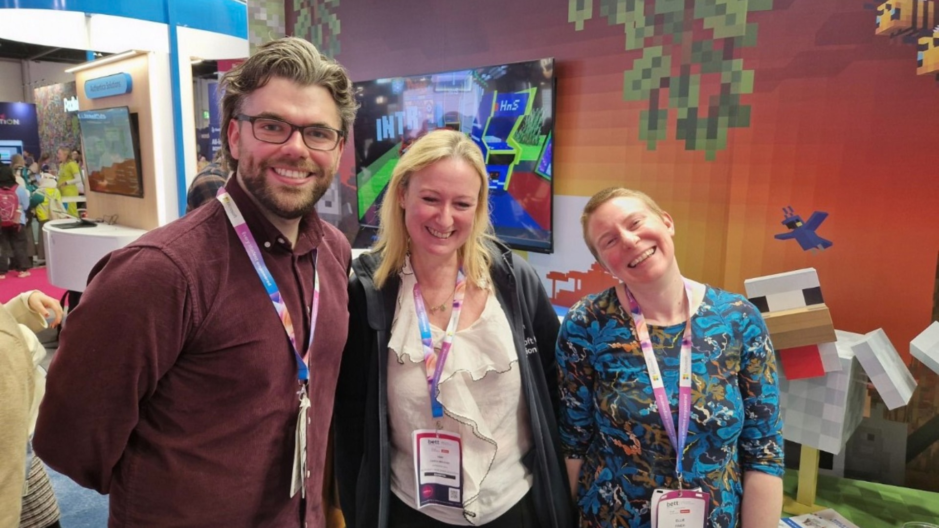 Three lanyard-wearing attendees, one man and two women, smile together in front of a Minecraft Education booth decorated with pixelated artwork.