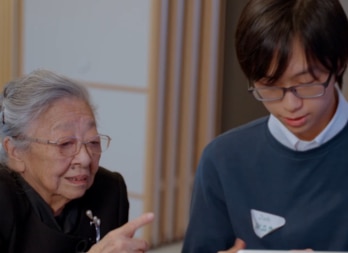 A man and an older woman sit together at a table, discussing and working on a laptop.