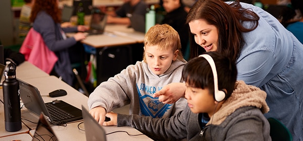 Two young students are using laptop computers. A teacher is providing instruction to them.