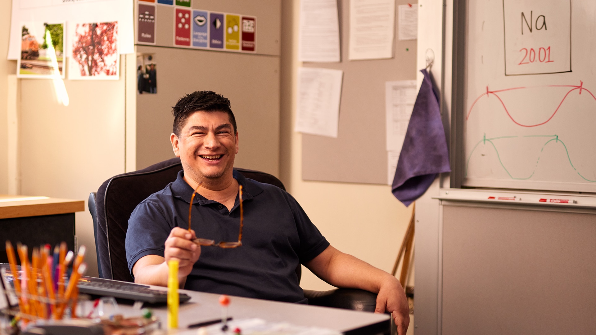 A smiling man holds glasses while sitting at a desk.