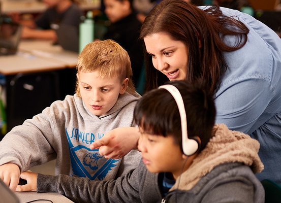 A smiling teacher leans in to assist two focused young students using a computer. One student wears headphones.