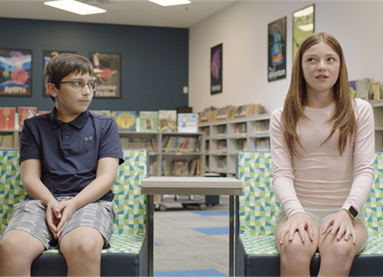 Two kids sit in a library on colorful chairs with a table between them. 